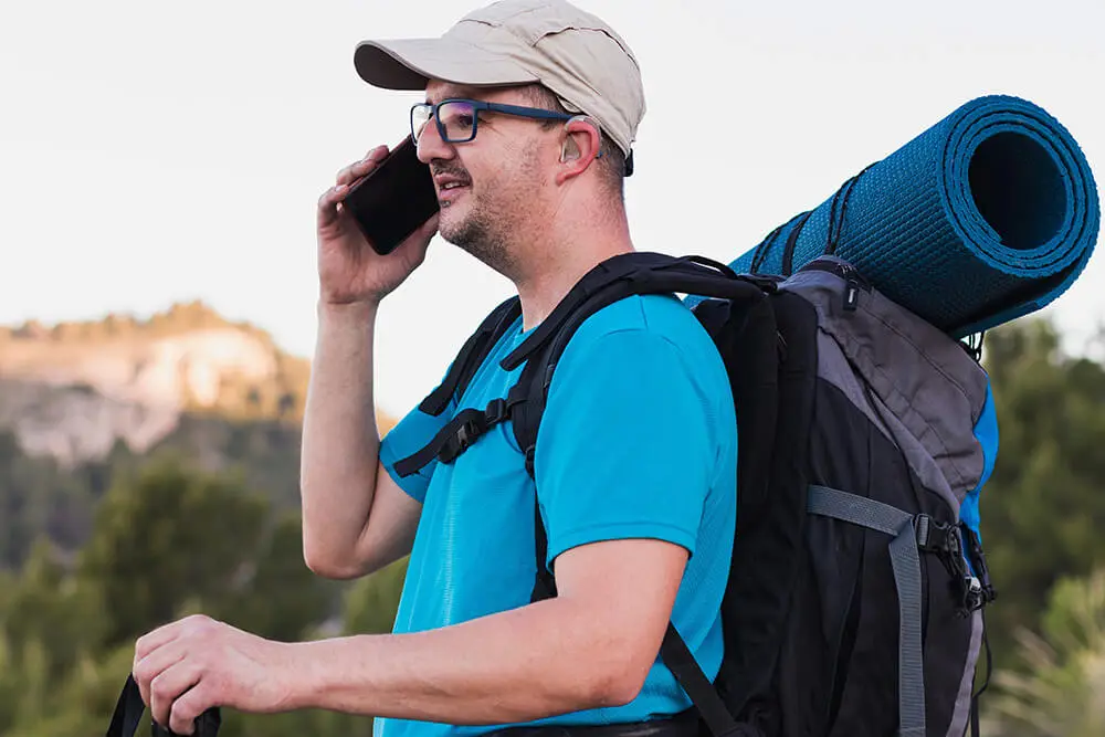 Man wearing glasses and a cap talks on a smartphone while hiking. He carries a backpack with a rolled-up mat, standing in a natural setting with blurred trees and hills in the background. Hes dressed in a blue shirt and sunglasses.