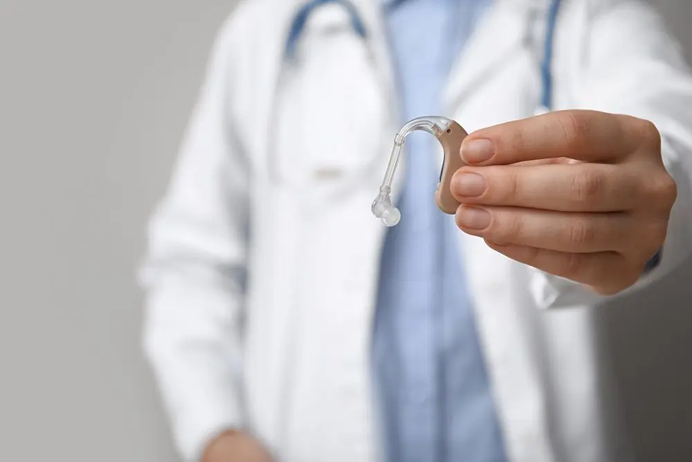 A person in a white coat holds a hearing aid close to the camera. The background is neutral, and the focus is on the hearing aid, which is beige with a clear earpiece.