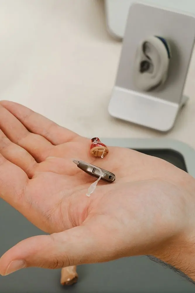 A hand holding two hearing aids of different styles, one red and the other silver. In the background, an ear model is visible on a stand.