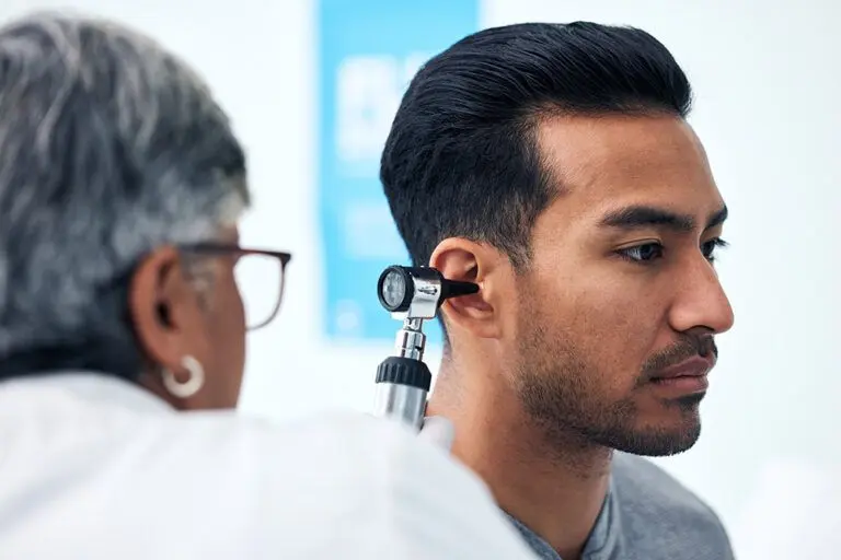 A healthcare professional examines a mans ear using an otoscope. The man has dark hair and a focused expression. The background is out of focus, with a blue poster partially visible.